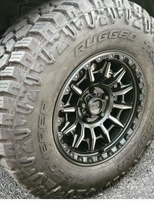 Close-up of a Cooper Discoverer Rugged Trek tire mounted on a black KMC wheel, showing its aggressive hybrid tread and alternating shoulder lugs for on- and off-road traction.