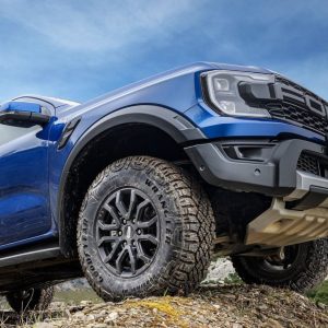 Close-up of a Goodyear Wrangler DuraTrac R/T tire mounted on a blue Ford Raptor, showing aggressive tread blocks and sidewall lugs designed for off-road terrain.