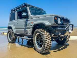 Suzuki Jimny equipped with BFGoodrich KM3 mud-terrain tires driving on wet beach sand, showcasing aggressive tread blocks and rugged off-road stance.