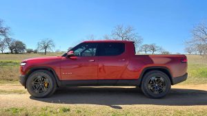 Side profile of a red Rivian R1T electric truck equipped with Continental TerrainContact H/T tires, parked on a dirt road under clear blue skies.