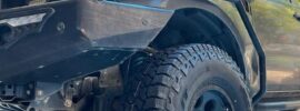 Close-up low-angle view of a lifted 4x4 truck fitted with Falken Wildpeak AT3W all-terrain tires, showing aggressive tread pattern and heavy-duty front bumper under a sunlit canopy.