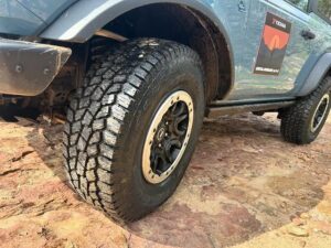 Close-up of a Yokohama Geolandar AT4 all-terrain tire mounted on a rugged SUV, parked on rocky terrain, showing aggressive tread pattern and black alloy wheel.